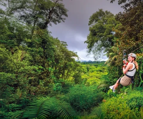Un viaggiatore che scende con una teleferica attraverso la fitta chioma della foresta pluviale costaricana, sospeso sopra una vegetazione rigogliosa con le montagne in lontananza.