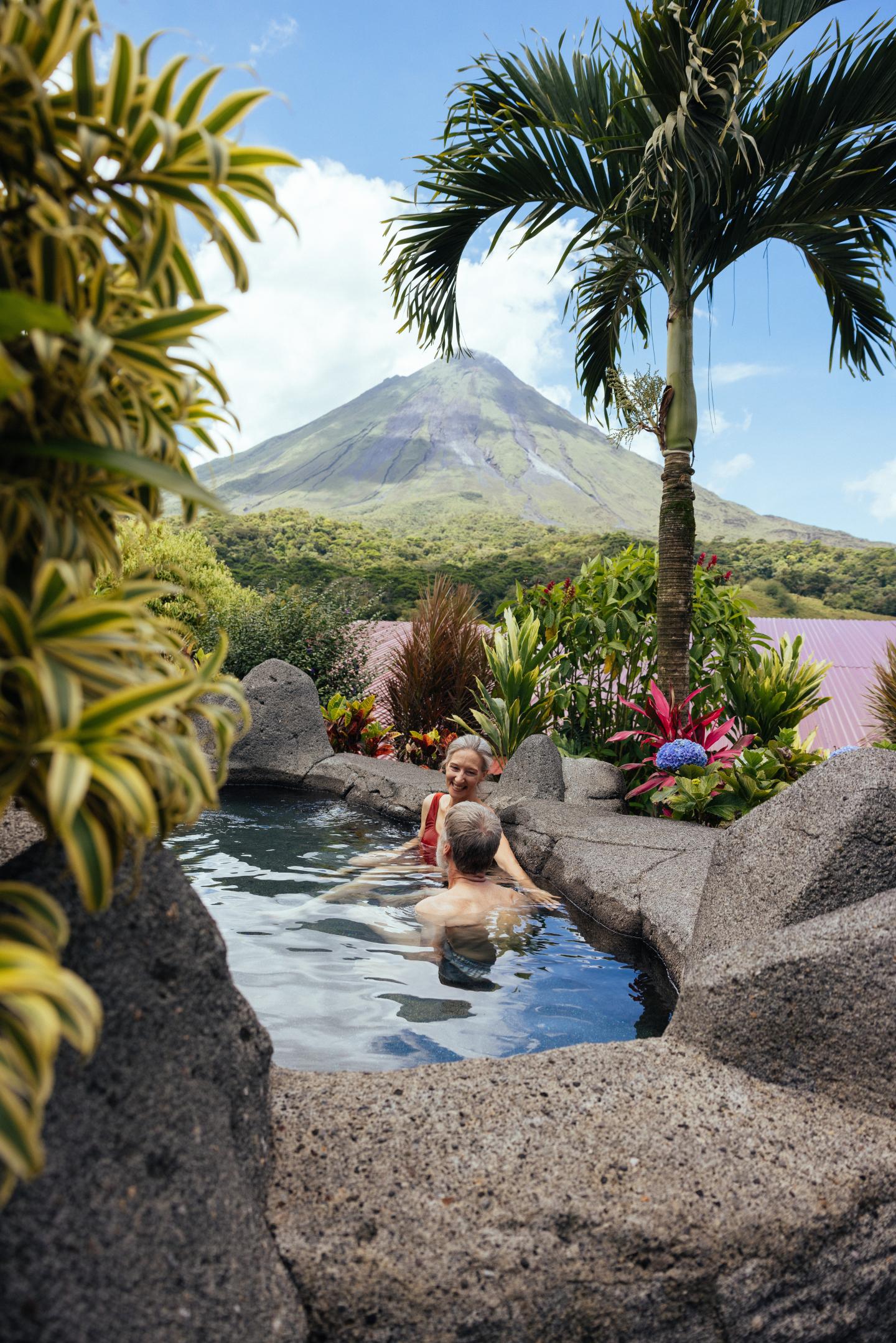 Due persone si rilassano in una vasca termale in pietra circondata da piante tropicali, con il vulcano Arenal sullo sfondo.